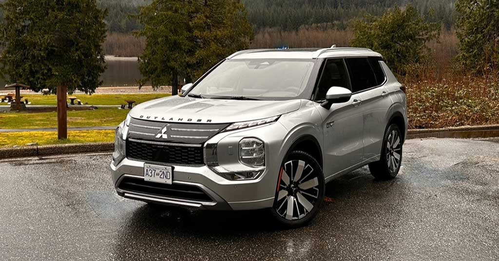 2026-Outlander-PHEV - West-Central Cross Roads Front-three-quarter view of a silver Mitsubishi Outlander SUV parked on a wet pavement with trees and a lake in the background, daytime outdoor scene.