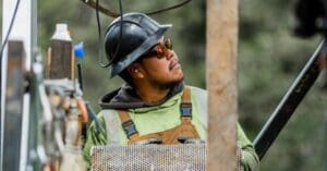 Construction worker wearing a hard hat and safety glasses, inspecting a perforated metal panel on a construction site.